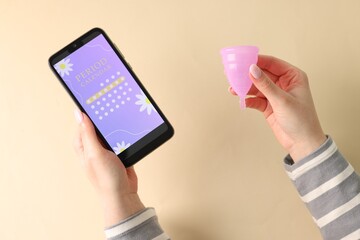 Menstruation. Woman holding smartphone with period calendar application and menstrual cup on beige...