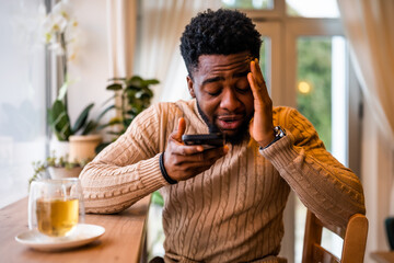 A young black man sits at the bar, casually dressed. He is using smartphone and recording voice message.