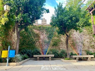 Rainbow Yarn Art and Ivy Wall in Spanish Courtyard. Blue and Yellow Recycling Bins near Green Wall and Tree. Recycling Concept.