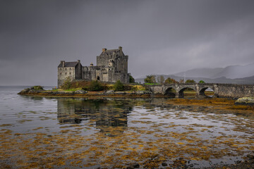 Eilean Donan Castle stands majestically amid autumn hues, its reflection shimmering in calm waters. Misty Highlands and golden trees create a scene of timeless beauty.