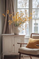 A bouquet of yellow flowers in a white vase sits on a white cabinet next to a comfy chair. Soft, natural light fills the room.