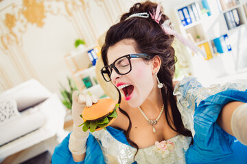 Elegant woman in historical costume enthusiastically eating a modern hamburger in a stylish and...