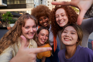 Happy selfie of a multiracial group of female friends gesturing a frame with fingers to camera,...