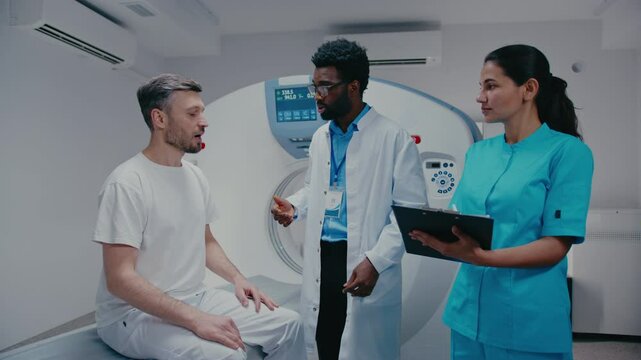 Male patient sitting on MRI table explaining symptoms while doctor listening closely. Placing his hand on shoulder. Female nurse in scrubs reviewing notes on clipboard. Noting important information.