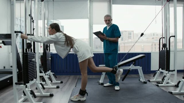 Side view of girl performing glute kickbacks on cable machine during physical therapy with senior male doctor controlling process and taking notes in gym