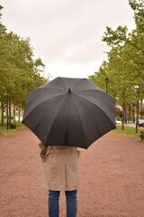 Rainy weather, man in trench coat standing in park with black umbrella