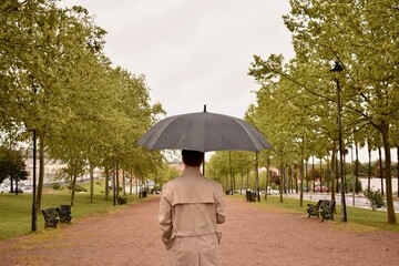 Rainy weather, man in trench coat standing in park with black umbrella