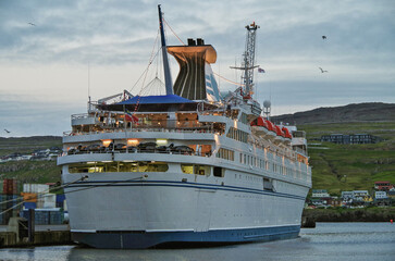 Naklejka premium Classic expedition cruiseship cruise ship liner Delphin docked at pier in Thorshavn, Faroe Islands with cliffs and fjord landscape in background and city skyline during Arctic Circle summer cruising