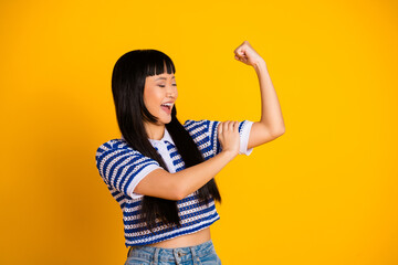 Cheerful young woman flexing arm to show strength against a vibrant yellow background wearing a...
