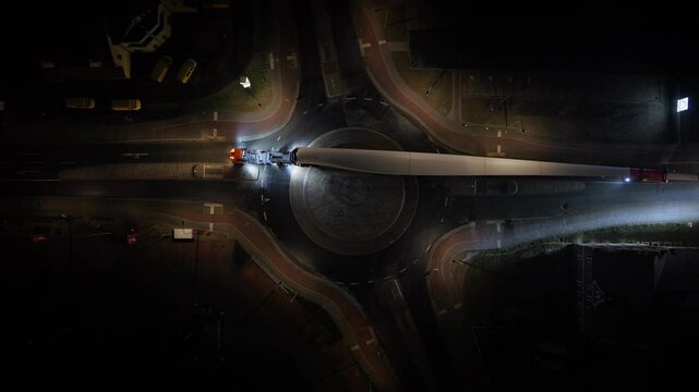 Top-down night shot of a truck transporting a massive wind turbine blade through a roundabout, headlights illuminating the road as it slowly turns right.