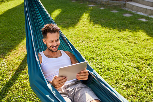 Handsome man in hammock using digital tablet relaxing in backyard