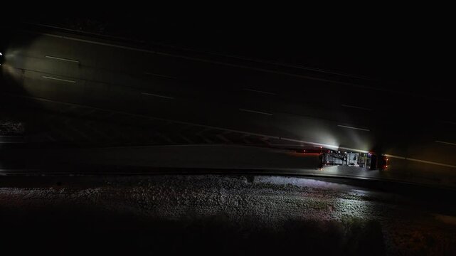 Top-down night view of a wind turbine blade convoy joining the highway, guided by escort vehicles with bright lights around the oversized load.