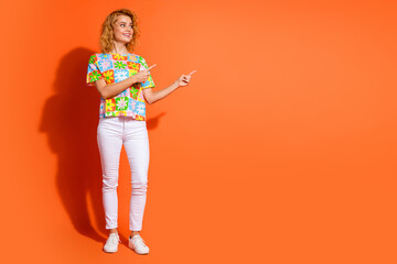 Confident young woman gestures while standing against vibrant orange background, wearing stylish floral top and white pants