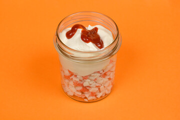 A top-down shot of a clear glass jar filled with layers of diced surimi sticks, cream, mayonnaise and a dollop of ketchup on top, suggesting the preparation of a surimi stick spread, against a vibrant