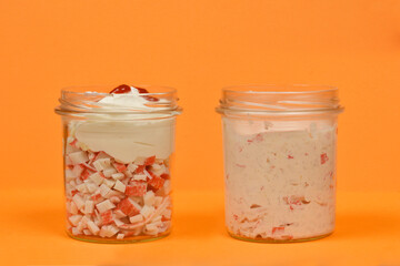 Two clear glass jars against an orange background, one with layered surimi sticks, cream, and a dollop of ketchup, and the other filled with a blended surimi spread