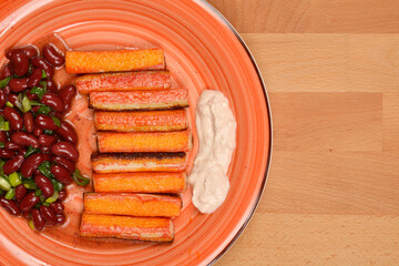 A top-down view of a plate featuring a row of grilled surimi sticks, a dollop of creamy sauce, and a side of red bean salad