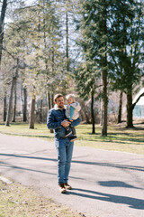 Obraz premium Smiling dad with a little girl in his arms walks along an asphalt road in a coniferous forest