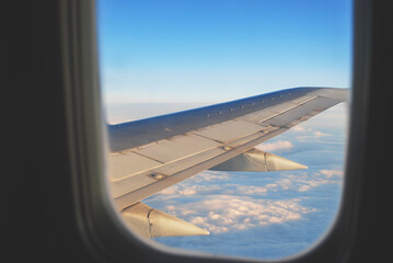 Airplane wing view from passenger window with soft clouds below and clear blue sky during flight, concept travel, freedom, aviation