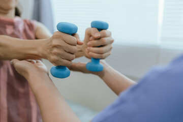 Senior woman exercising with dumbbells during physical therapy in hospital. Physiotherapist helps...
