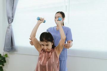 Senior woman exercising with dumbbells during physical therapy in hospital. Physiotherapist helps...