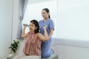 Senior woman exercising with dumbbells during physical therapy in hospital. Physiotherapist helps...