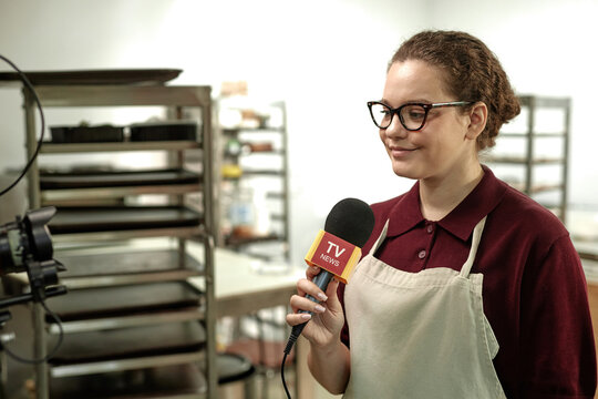 Chest up shot of cheerful young female baker in apron giving interview to journalists holding microphone captured in pastry shop kitchen, copy space