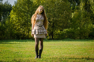 Young woman with long red hair walks through sunlit park. Wearing beige dress and boots, she...