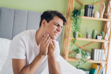 Young man experiencing discomfort while holding his jaw in a well-lit bedroom with plants and cozy...