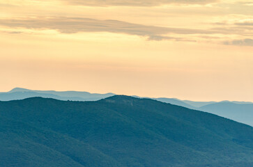 Layered mountain ridges under soft, golden dusk light create serene atmosphere. Sky filled with delicate clouds adds tranquility to scene. Calming blend of blue and amber hues