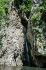 Obraz premium Slender waterfall descends through rocky gorge surrounded by lush greenery. Sunlight illuminates scene, highlighting texture of rocks. Water pools at base, forming serene reflections