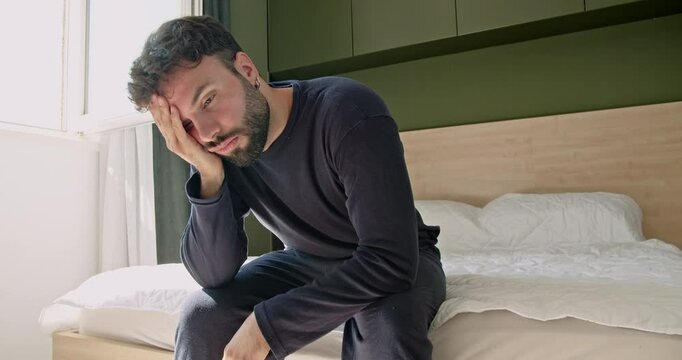 A young Caucasian man wakes up in his modern bedroom, sits on the bed, and stretches while yawning and rubbing his eyes.