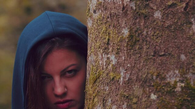 Beautiful witch with long red hair and dark blue cloak hiding and peeking around tree.