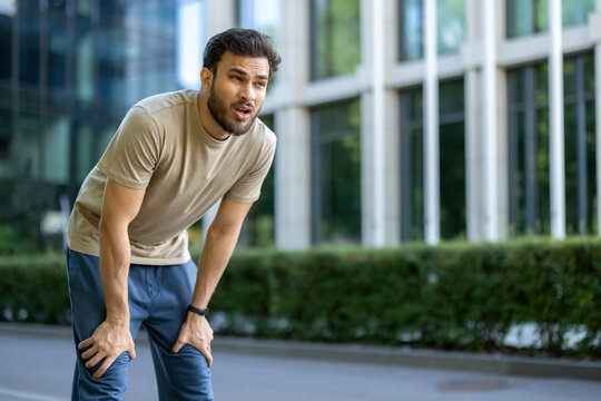 Tired young Indian man who is involved in running and sports, standing outside and resting, bent over, breathing heavily