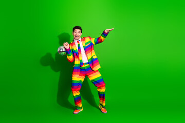 Handsome young man in a vibrant rainbow striped suit posing joyfully against a vivid green background while holding a disco ball.