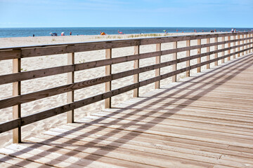 Obraz premium Wooden boardwalk casts shadows across its planks as it meanders beside peaceful sandy beach. In distance, colorful umbrellas dot shore, inviting tranquil seaside escape