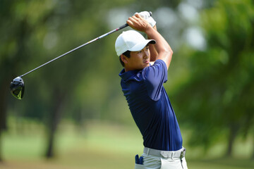Asian male golfer in blue shirt swinging club on golf course