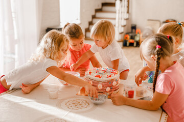 Group of cheerful kids decorating birthday cake together in bright home kitchen during celebration
