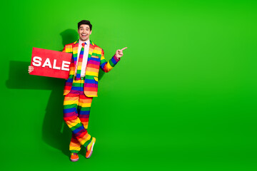 Young man in vibrant rainbow suit holding a sale sign on a green background, conveying a joyful message of fun, fashion, pride
