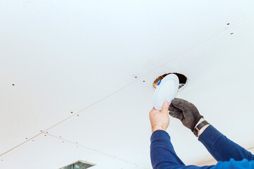 Electrician is fixing recessed light fixture in ceiling as home remodel in bright indoor environment.