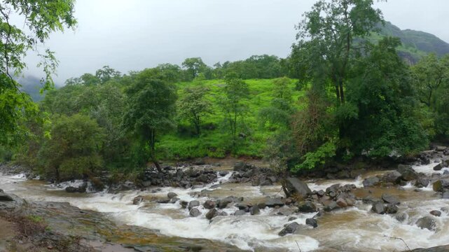 4K shot of river stream besides the hill with green forest and grass during the monsoon season and dark clouds in the sky as seen on the way towards Devkund waterfall in Raigad, Maharashtra, India.