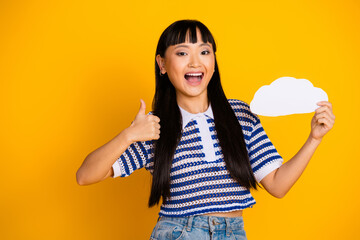 Cheerful young woman in casual attire holding a cloud cutout against a vibrant yellow background,...