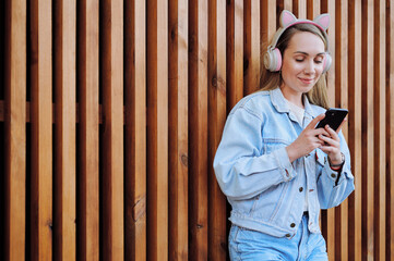 Stylish young woman in denim jacket and pink cat-ear headphones holding smartphone, leaning...