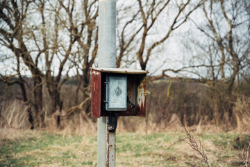 A rusted metal box with a small window sits on a pole in a field