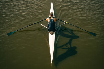 Top view of a person rowing in a single scull boat on calm water, casting a shadow, showcasing balance and athleticism.