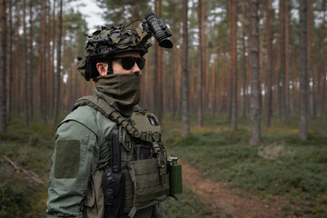 Soldier in full tactical gear, helmet with NVG mount, and sunglasses, patrolling a dense pine...