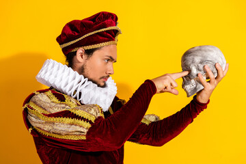 Young nobleman in historical costume pointing at human skull against a yellow background, to be or...