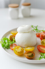 Buratta on a gray plate with cherry tomatoes and herbs on a white table. Traditional Italian food concept. Vertical orientation. Selective focus.