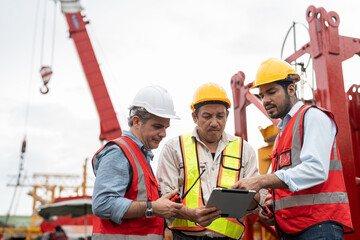 Team of construction civil engineers or workers wearing safety gear examine project plans on digital tablet at construction site.