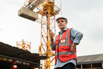 Construction site supervisor or project manager wearing a white hard hat and safety vest using a walkie-talkie standing near red tower crane equipment.