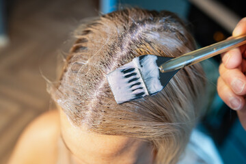 Home hair dyeing: Close-up of applying dye with a brush to regrown roots of blonde hair at home.
